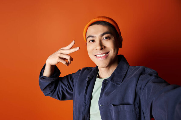 Stylish Asian man in a blue jacket and orange hat poses confidently against an orange background in a studio.