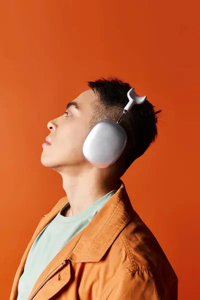 オレンジ色のスタジオの背景に対して彼の耳にヘッドフォンで音楽を聞くスタイリッシュな服装のハンサムなアジア人男性 — ストック写真