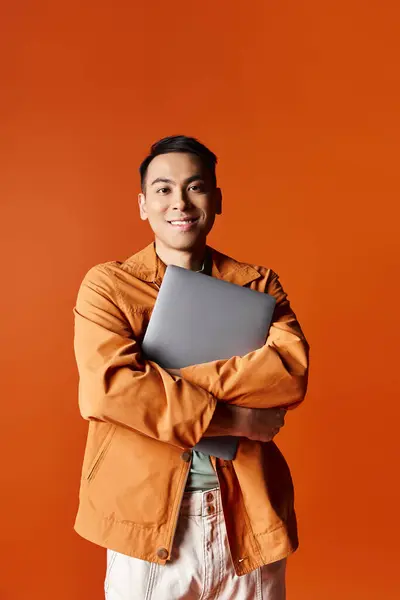 一个时髦的亚洲男人 双手交叉 自信地拿着笔记本电脑 背对着橙色的背景 — 图库照片