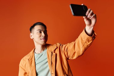 Yakışıklı Asyalı şık bir adam cep telefonuyla portakal rengi bir stüdyonun arka planında fotoğraf çekiyor..