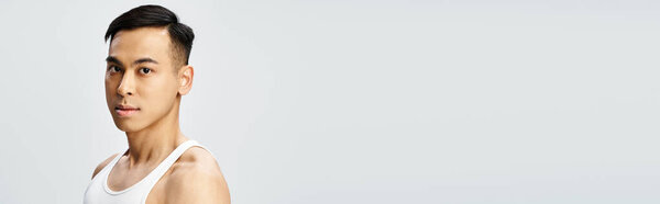 A handsome Asian man in a white tank top strikes a confident pose in a grey studio setting.