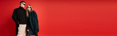 Şık ceketli, güneş gözlüklü, kırmızı arka planda poz veren genç bir çift.