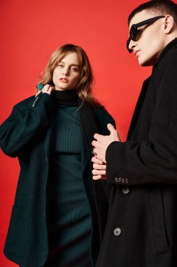 Zarif ceketli çekici, uzun saçlı kadın güneş gözlüklü erkek arkadaşının yanındaki kameraya bakıyor.