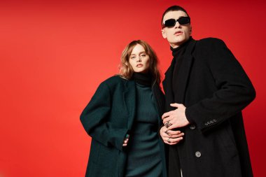 Zarif ceketli çekici, uzun saçlı kadın güneş gözlüklü erkek arkadaşının yanındaki kameraya bakıyor.