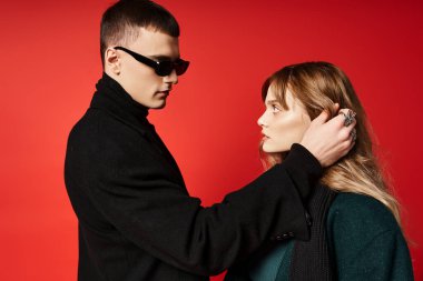 Güzel bir genç kadın ve güneş gözlüklü erkek arkadaşı kırmızı arka planda birbirlerine bakıyorlar.