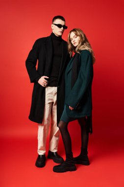 Zarif ceketli güzel, uzun saçlı kadın güneş gözlüklü erkek arkadaşının yanındaki kameraya bakıyor.