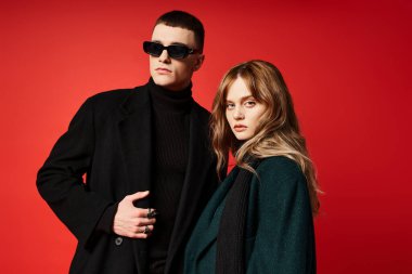 Zarif ceketli güzel, uzun saçlı kadın güneş gözlüklü erkek arkadaşının yanındaki kameraya bakıyor.