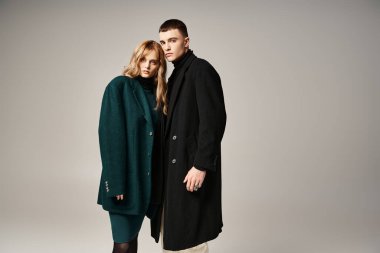 Gri arka planda birlikte poz verip kameraya bakan şık ceketli sofistike bir çift.