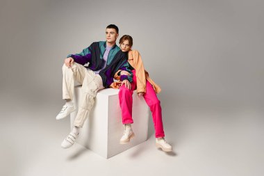 alluring woman in fashionable bomber looking at camera next to her handsome boyfriend on white cube clipart