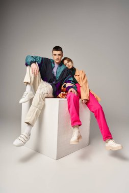 Canlı stil bombardıman uçaklarıyla beyaz kübün üzerindeki gri arkaplanda kameraya bakan şık çifti seviyorlar.