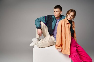 alluring woman in fashionable bomber looking at camera next to her handsome boyfriend on white cube clipart