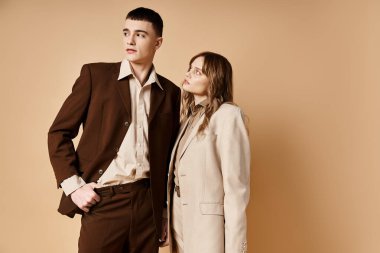 Tarz sahibi takım elbiseli, sevimli bir çift birlikte poz veriyor ve pastel arka plana bakıyor.