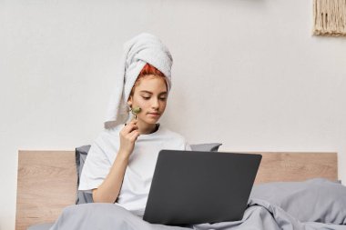 Ev elbiseleri içindeki kızıl saçlı homo, laptopla yatakta dinlenirken yüz silindiri kullanıyor.