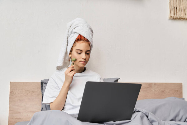 привлекательный рыжеволосый педик в домашней одежде с помощью ролик для лица во время отдыха на кровати с ноутбуком