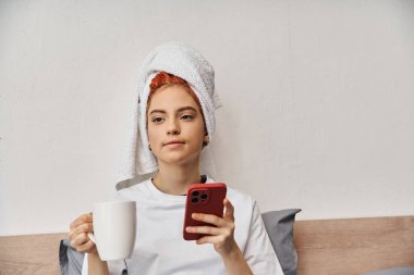 pozitif rahatlatıcı ev elbiseli tuhaf insan saç havlusuyla telefon kullanıyor ve yatakta çay içiyor.