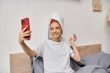 cheerful beautiful queer person with hair towel in homewear taking selfies while relaxing in bed clipart