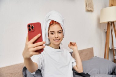 cheerful beautiful queer person with hair towel in homewear taking selfies while relaxing in bed clipart