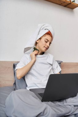 Neşeli, güzel, tuhaf saçlı, laptopta film izleyen ve yatakta gua sha kullanan biri.