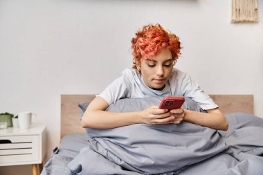 Uykucu, güzel, ev elbiseli, kızıl saçlı, yatakta oturan ve akıllı telefonunu kullanan tuhaf insan.