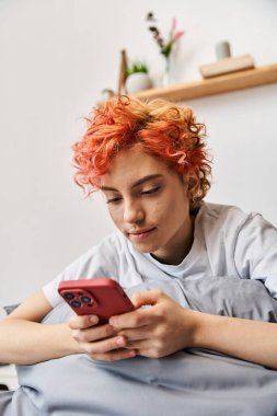 Uykucu, güzel, ev elbiseli, kızıl saçlı, yatakta oturan ve akıllı telefonunu kullanan tuhaf insan.