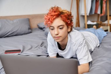Keyifli, çekici, rahat ev elbiseli, yatakta uzanan ve internette sörf yapan homo.