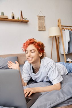 good looking red haired cheerful queer person in homewear having video call while lying in bed clipart