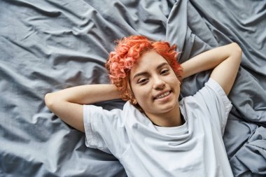 cheerful appealing queer person with red hair lying on her bed and looking at camera, leisure time clipart