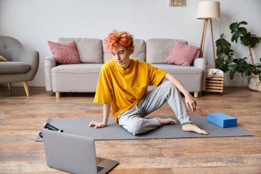 Sarı tişörtlü, yoga minderinde oturan ve egzersiz yapmadan önce dizüstü bilgisayara bakan güzel tuhaf insan.