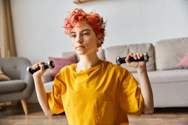Canlı spor kıyafetleri içinde, evde halterlerle egzersiz yapan çekici, tuhaf bir genç.