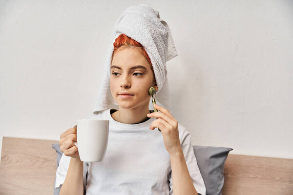 привлекательный странный человек с полотенцем для волос с помощью ролик лица и держа чашку чая в руке в то время как дома
