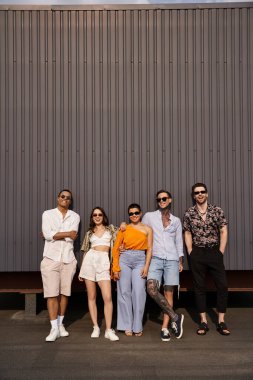 Bir grup neşeli çoklu-ırkçı arkadaş, günlük kıyafetleriyle duvarda poz veriyor ve kameraya gülümsüyor.