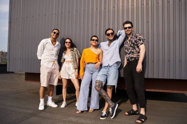 Duvarda poz verip kameraya gülümseyen neşeli, çok kültürlü arkadaşlar.