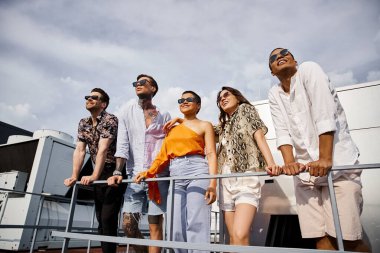 Neşeli, güzel, ırklar arası, sıradan giysiler içinde çatıda poz verip başka tarafa bakan arkadaşlar.