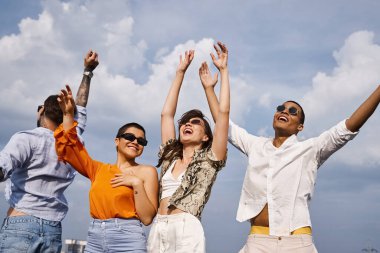 joyful appealing multiracial friends in vibrant clothing posing actively on rooftop together clipart