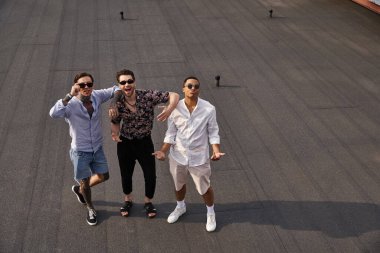 Çok kültürlü, neşeli, şık güneş gözlüklü erkek arkadaşlar çatıda poz verip kameraya bakıyorlar.