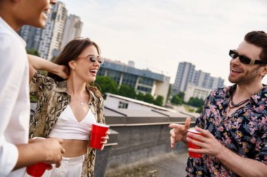 interracial cheerful friends in vibrant attires drinking from red cups at rooftop party together clipart