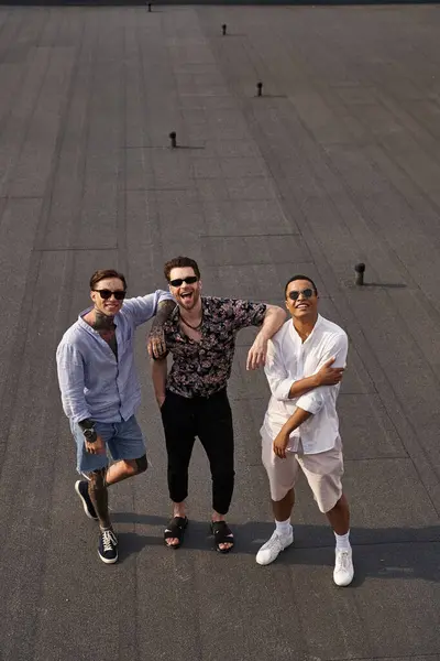 three diverse cheerful male friends with sunglasses posing on rooftop and looking at camera
