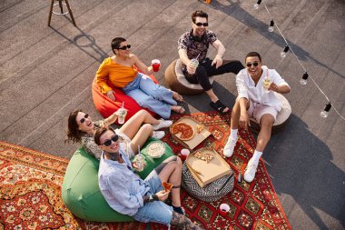 Çatıdaki partide pizza yiyip içen ve kameraya gülümseyen çeşitli neşeli arkadaşların üst görüntüsü.