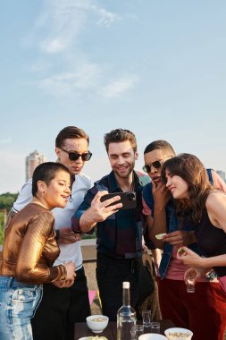 Çok kültürlü genç insanlar, gündelik giysiler içinde çatıdaki partide akıllı telefondan fotoğraf izliyorlar.