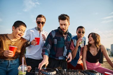 interracial joyous people in vibrant outfits partying together next to handsome DJ at rooftop party clipart