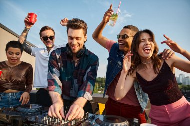 joyful good looking interracial friends in urban attires partying together with handsome DJ at party clipart