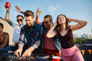 Canlı giysiler içinde neşeli çok kültürlü insanlar çatı partisinde çekici DJ 'in yanında parti yapıyorlar.