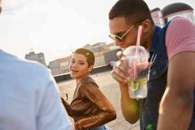 Canlı giyinmiş neşeli kısa saçlı kadın partide farklı arkadaşlarının yanında kameraya gülümsüyor.