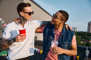 good looking joyous interracial men holding cocktails and looking at each other at rooftop party clipart