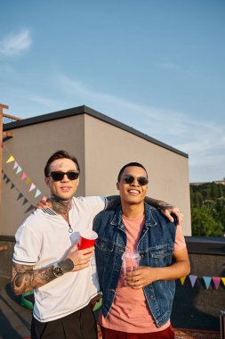 jolly multicultural men in vibrant urban attires holding cocktails and looking at camera at party clipart