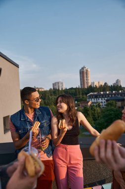 jolly multiracial friends in vibrant outfits enjoying delicious hot dogs on rooftop at party clipart