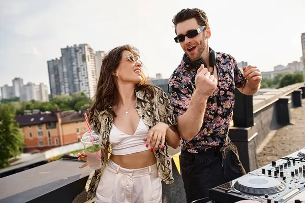 joyous attractive woman with cocktail dancing with handsome joyous DJ to music at rooftop party