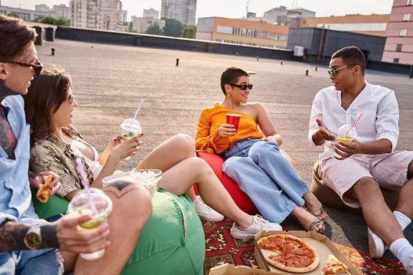 joyous multiracial friends with sunglasses sitting on rooftop at party with cocktails and pizza