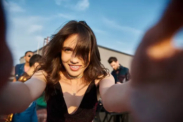 focus on joyful beautiful woman looking at camera at rooftop party with her diverse blurred friends