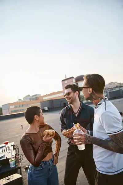 joyful good looking people with sunglasses in vivid outfits enjoying hot dogs at rooftop party